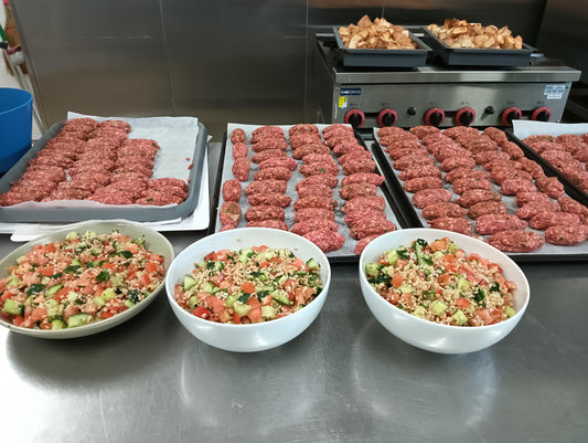 Multiple trays with beautiful chef made lamb kofta, next to white bowels of fresh Greek salad.